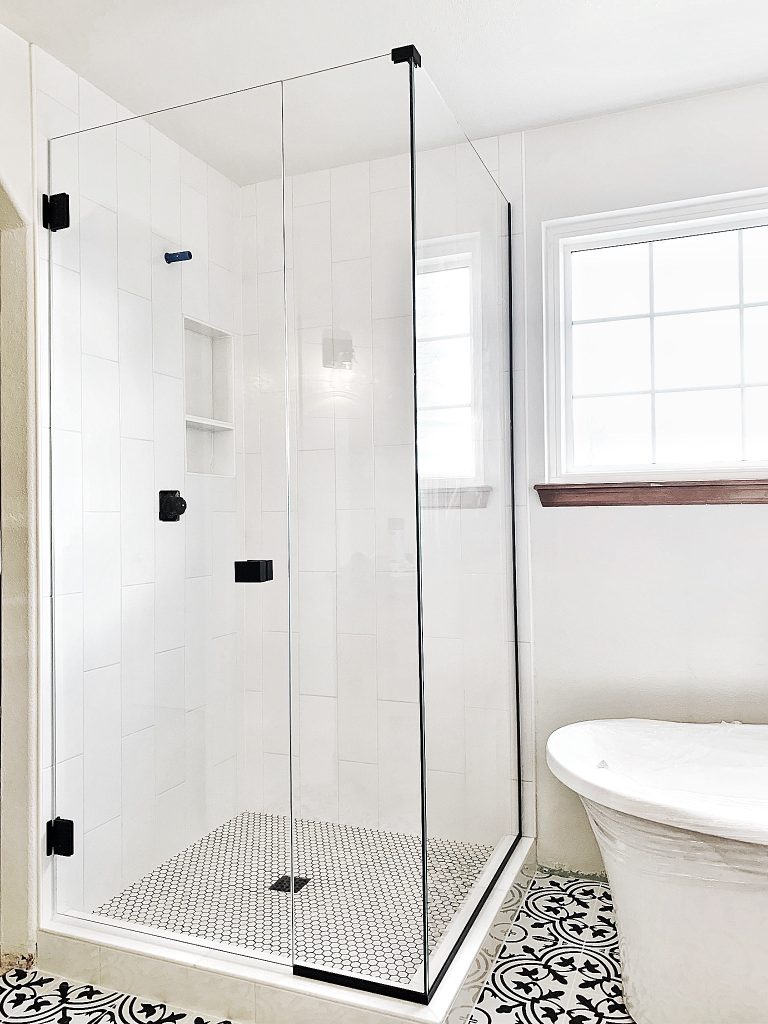 A modern bathroom with a glass shower enclosure by a Glass Company Austin, white tiled walls, black hardware, and a white freestanding bathtub next to patterned black and white floor tiles.