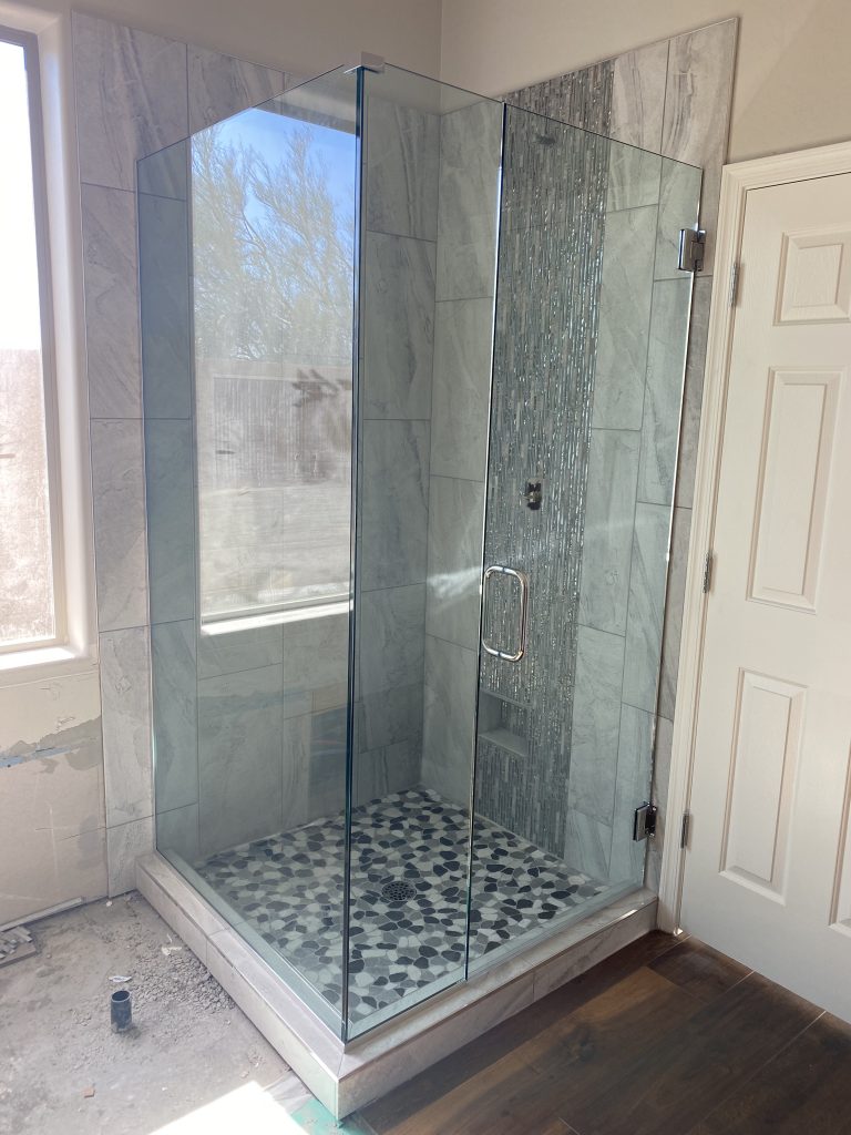 A modern, glass-enclosed shower with marble tile walls, a vertical mosaic accent, and a pebble-patterned floor. Natural light highlights the clean, contemporary design of this Custom Glass Shower.