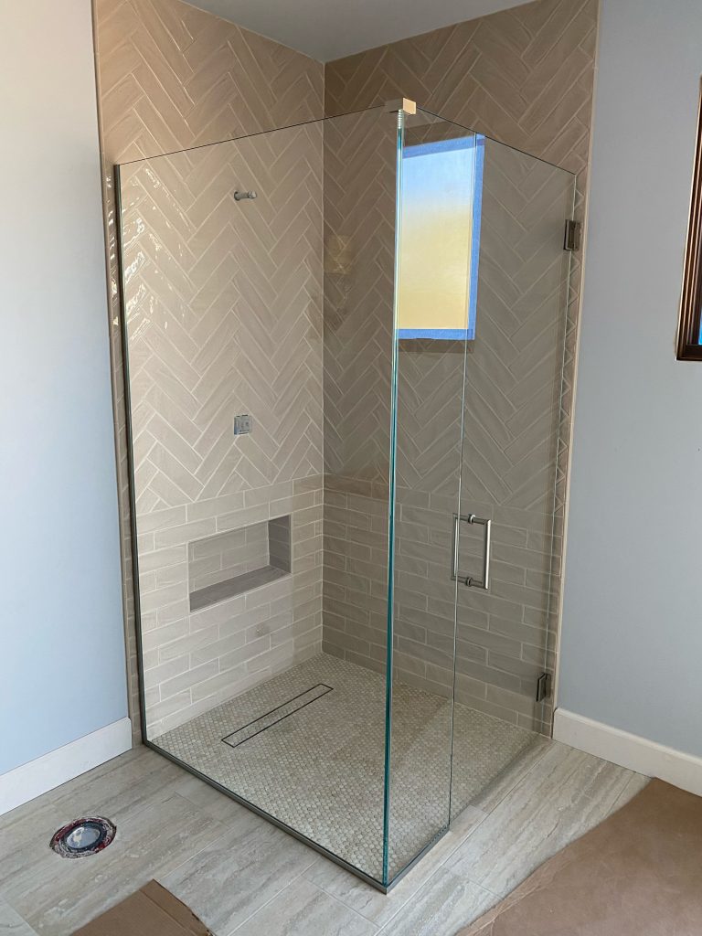 Modern glass shower with beige herringbone wall tiles, a built-in shelf, and a frameless glass door. The floor is light tile with a linear drain, designed by Glass Company Austin to complete your custom glass shower retreat.