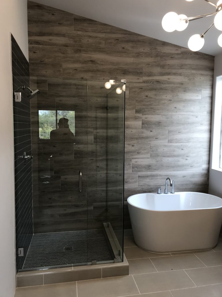 Modern bathroom with a custom glass shower enclosure, soaking bathtub, and wood-look tile walls. Natural light fills the room; an overhead fixture with round bulbs is visible. A person’s reflection appears in the shower glass.