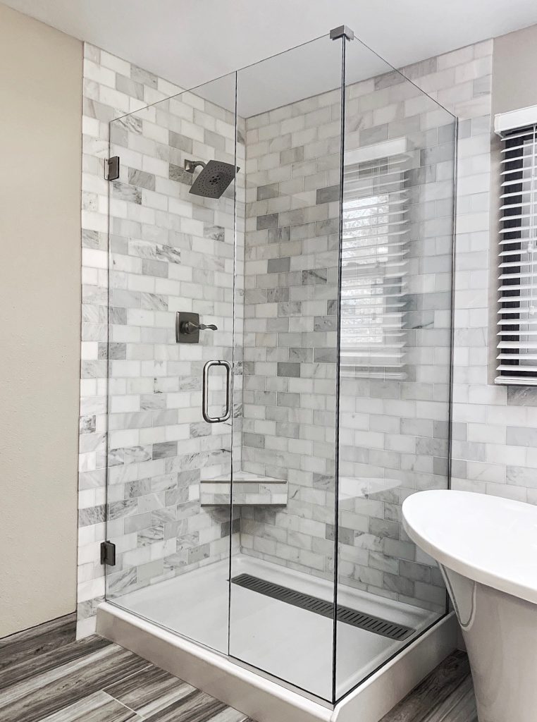 A modern frameless glass shower enclosure with a metal handle, wall-mounted showerhead, and built-in corner shelf. The walls showcase white and gray marble tiles, while the floor features gray wood-look tiles. A white bathtub is nearby.