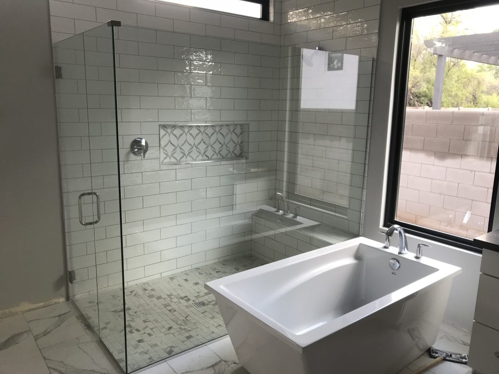 Modern bathroom with a glass-enclosed shower featuring white subway tiles, a built-in niche with a geometric pattern, and a sleek white freestanding bathtub on a tiled floor. Custom Glass Shower Enclosures add elegance to this serene space.