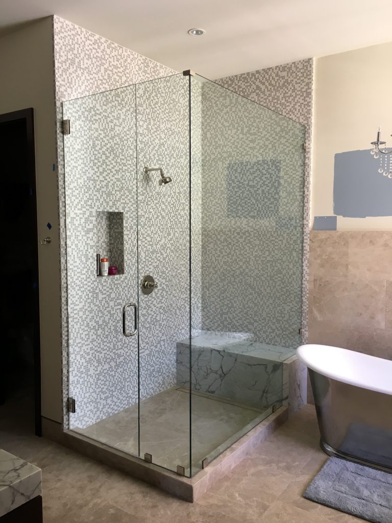 A modern bathroom featuring a frameless glass shower with mosaic tile walls, a built-in bench, a small recessed shelf, and a freestanding bathtub on beige marble flooring. A patch of blue paint accents the wall.