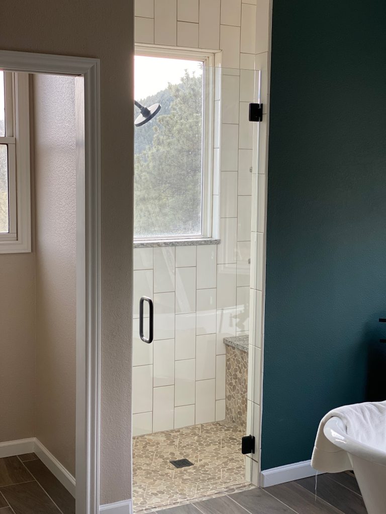 A modern bathroom features a Custom Glass Shower Enclosure, large vertical white tiles, a built-in bench with stone tile, and a dark green wall. Part of a tub is visible, and the space feels open with natural light illuminating the room.