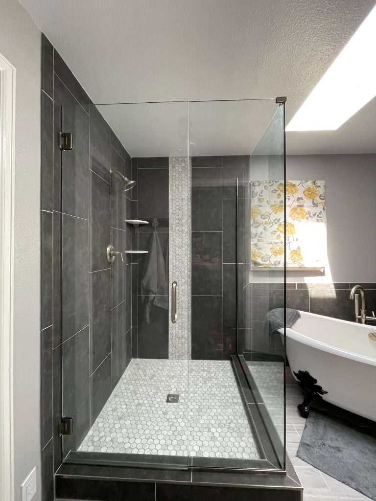 A modern bathroom with a frameless glass shower featuring dark tiles and a vertical mosaic accent, next to a freestanding white bathtub and natural sunlight streaming in.