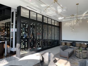 A modern lounge featuring a large glass wine cellar wall filled with wine bottles, gray seating, marble floors, and hanging globe light fixtures, with sunlight streaming in. The design echoes the elegance of Custom Glass Shower Enclosures. A modern lounge featuring a large glass wine cellar wall filled with wine bottles, gray seating, marble floors, and hanging globe light fixtures, with sunlight streaming in. The design echoes the elegance of Custom Glass Shower Enclosures.