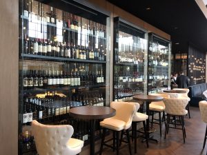 Modern wine bar interior with glass walls from a top Glass Company in Austin, showcasing shelves of wine bottles. Round tables are surrounded by cream-colored, tufted chairs on a wooden floor, with a few people visible in the background. Modern wine bar interior with glass walls from a top Glass Company in Austin, showcasing shelves of wine bottles. Round tables are surrounded by cream-colored, tufted chairs on a wooden floor, with a few people visible in the background.