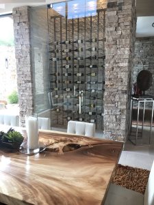 Modern dining area with a natural wood table, white chairs, and stone walls. Behind glass doors is a wine cellar with neatly arranged bottles. Decorative candles and plants adorn the table. Modern dining area with a natural wood table, white chairs, and stone walls. Behind glass doors is a wine cellar with neatly arranged bottles. Decorative candles and plants adorn the table.