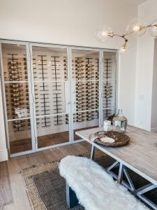 Modern dining room with a light wood table, fur-covered bench, wicker tray decor, and a glass-enclosed wine cellar displaying rows of wine bottles on metal racks. Contemporary chandelier hangs above. Modern dining room with a light wood table, fur-covered bench, wicker tray decor, and a glass-enclosed wine cellar displaying rows of wine bottles on metal racks. Contemporary chandelier hangs above.