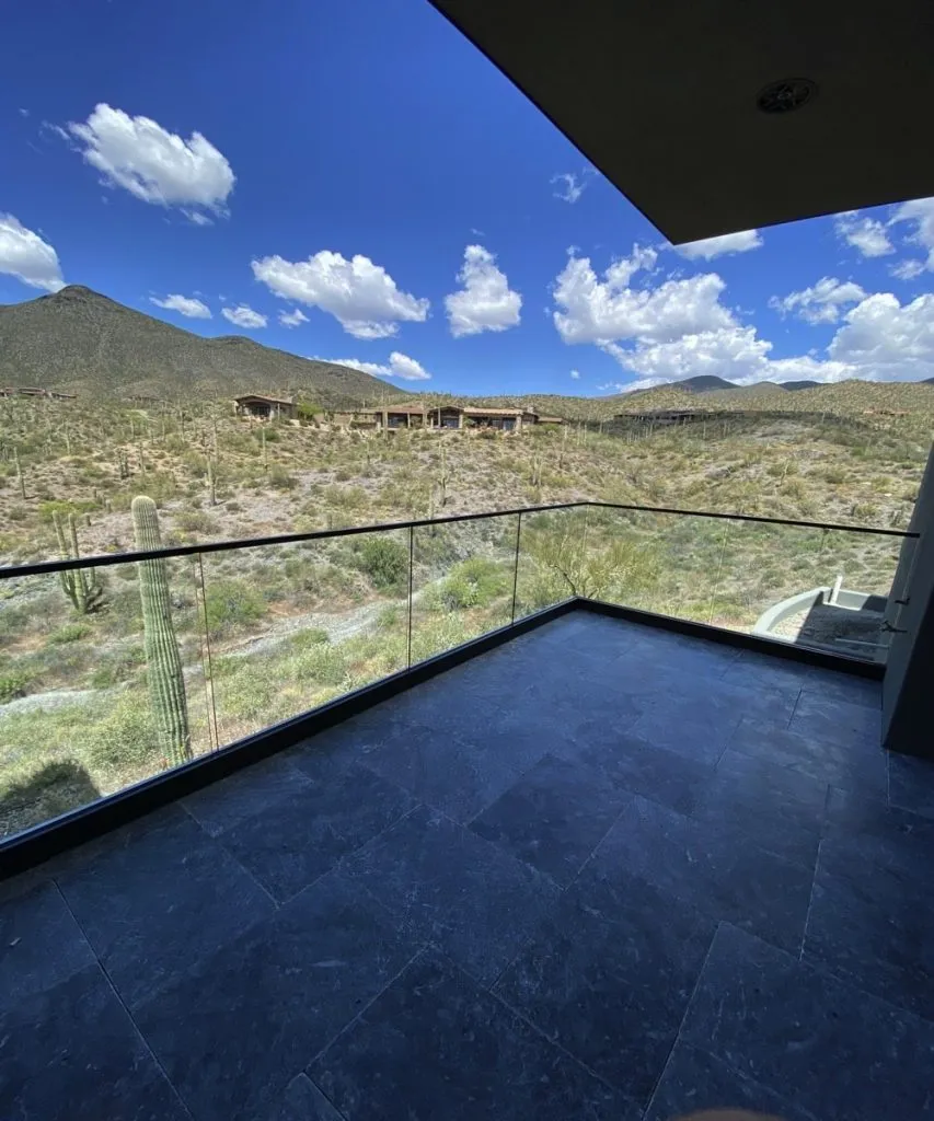 A modern balcony with a glass railing by Glass Company Austin overlooks a desert landscape with cacti, hills, and a blue sky filled with scattered white clouds.