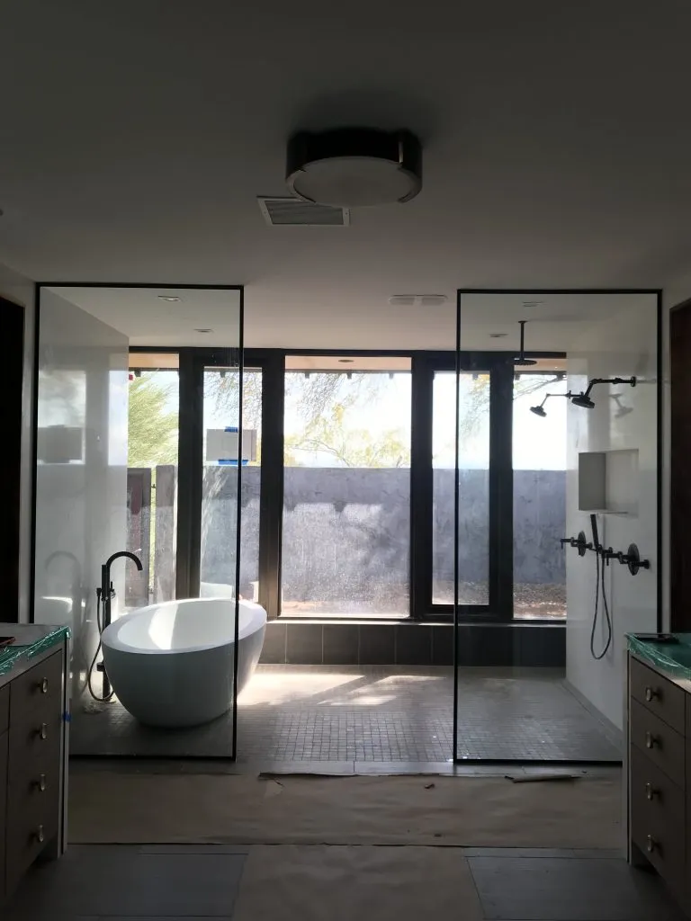A modern bathroom features a freestanding bathtub on the left and an Instagram worthy shower design on the right, separated by glass partitions. Floor-to-ceiling windows at the back let natural light fill the space.